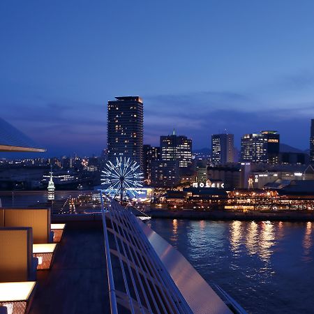 فندق كوبي ميريكين بارك أورينتال كوبه المظهر الخارجي الصورة The photo displays a panoramic view of a waterfront cityscape at twilight. You can see a vibrant skyline with towering buildings lit up against the evening sky. In the foreground, there are modern architectural elements, possibly part of a hotel or r