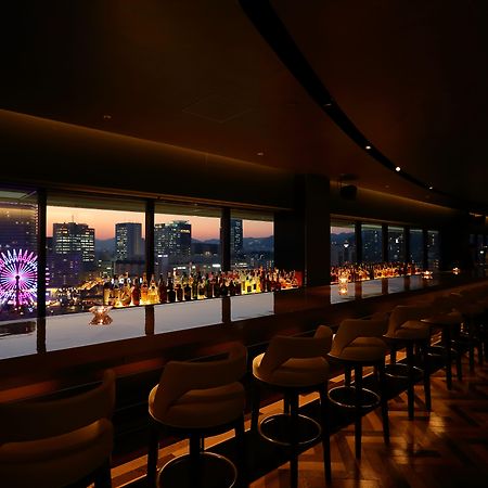فندق كوبي ميريكين بارك أورينتال كوبه المظهر الخارجي الصورة The photo shows the interior of a stylish bar or lounge with a panoramic view of a city skyline at dusk. The lighting is dim, creating an intimate atmosphere. There are elegant bar stools lined up along a long counter, which features an array of illu