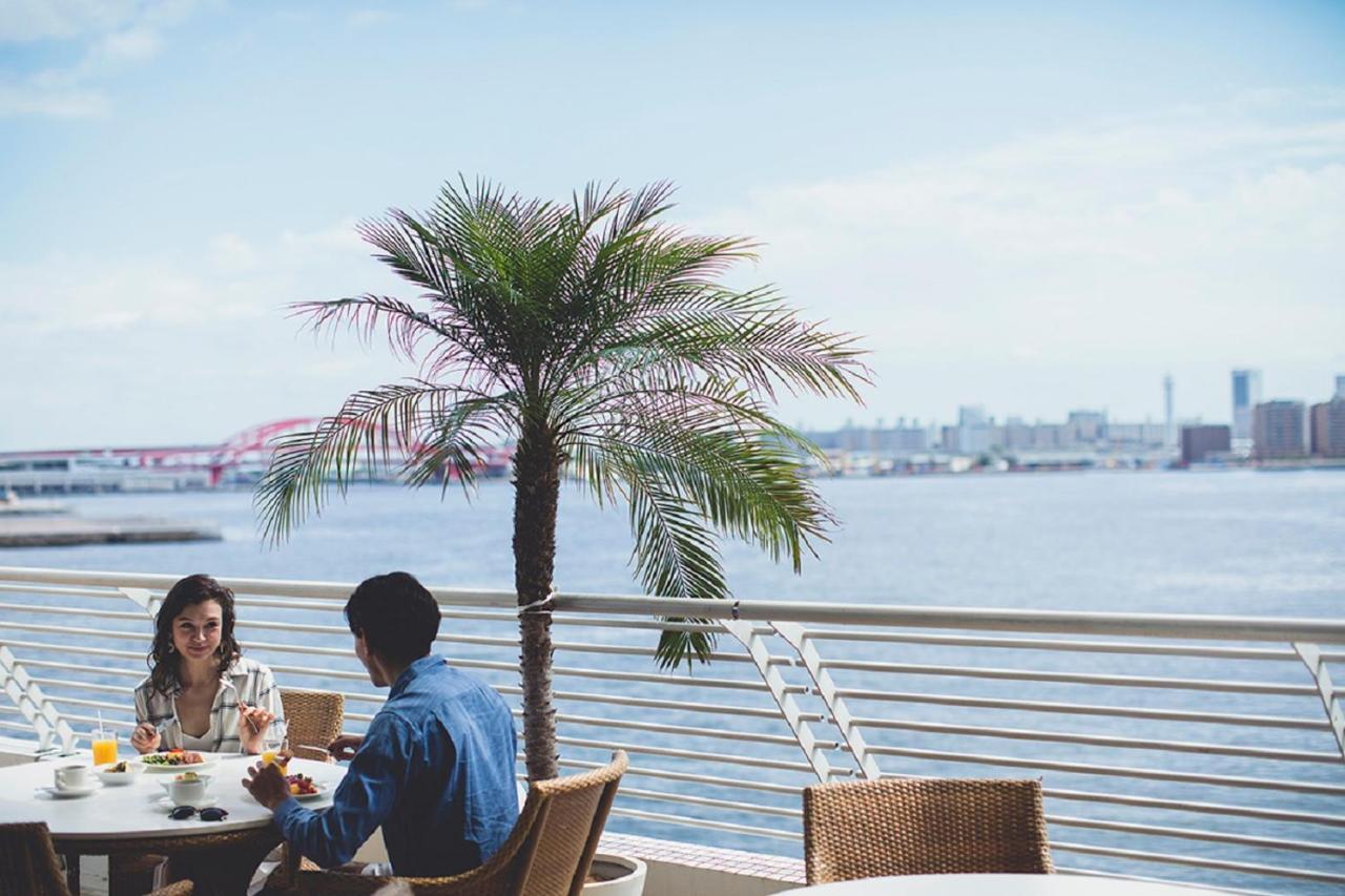 فندق كوبي ميريكين بارك أورينتال كوبه المظهر الخارجي الصورة The photo depicts a scenic outdoor dining setting by the water. In the foreground, there are two people sitting at a table with a stylish arrangement of food and drinks. A palm tree provides a tropical ambiance, standing prominently next to them. In 