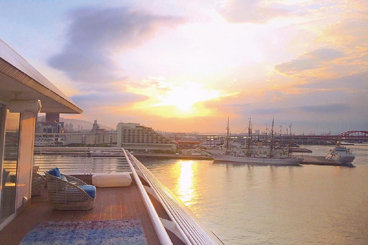 فندق كوبي ميريكين بارك أورينتال كوبه المظهر الخارجي الصورة The photo shows a serene waterfront scene with a stunning sunset. In the foreground, there is a deck area that appears to be part of a ship or a waterfront building, featuring a cozy seating arrangement. The deck has a soft rug and some cushions, sug