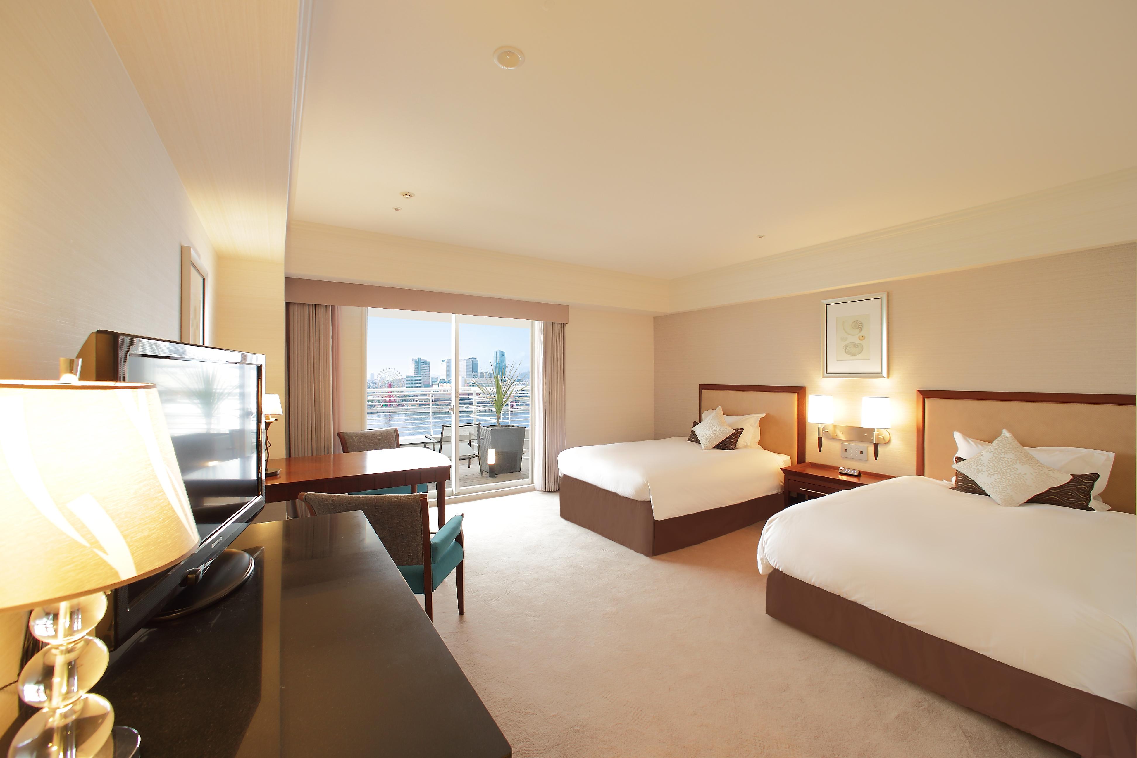فندق كوبي ميريكين بارك أورينتال كوبه المظهر الخارجي الصورة The photo shows a hotel room featuring two beds: one is a double bed while the other appears to be a twin bed. The room is well-lit, with large windows offering a view of the city. There is also a desk and a chair, along with a television on a dresse