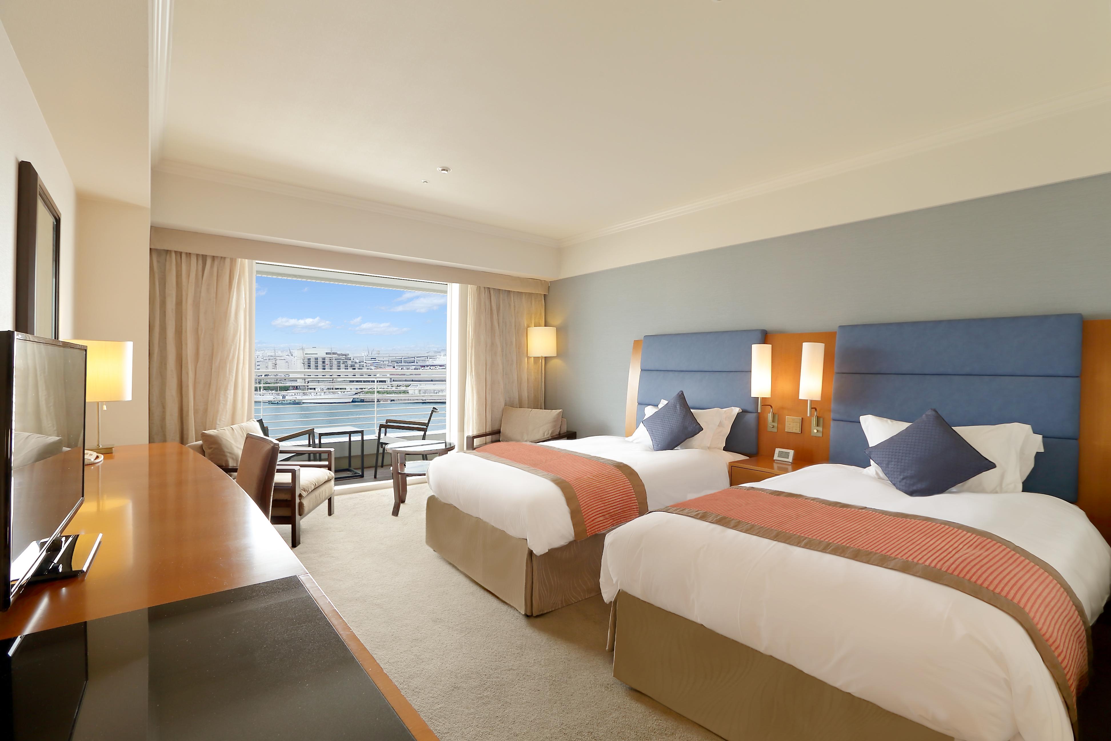 فندق كوبي ميريكين بارك أورينتال كوبه المظهر الخارجي الصورة The photo shows a hotel room that features two beds with white linens and orange accents. There is a wooden desk and chair positioned near the entrance, along with a modern chair and a small table next to the window. The room has large windows that a