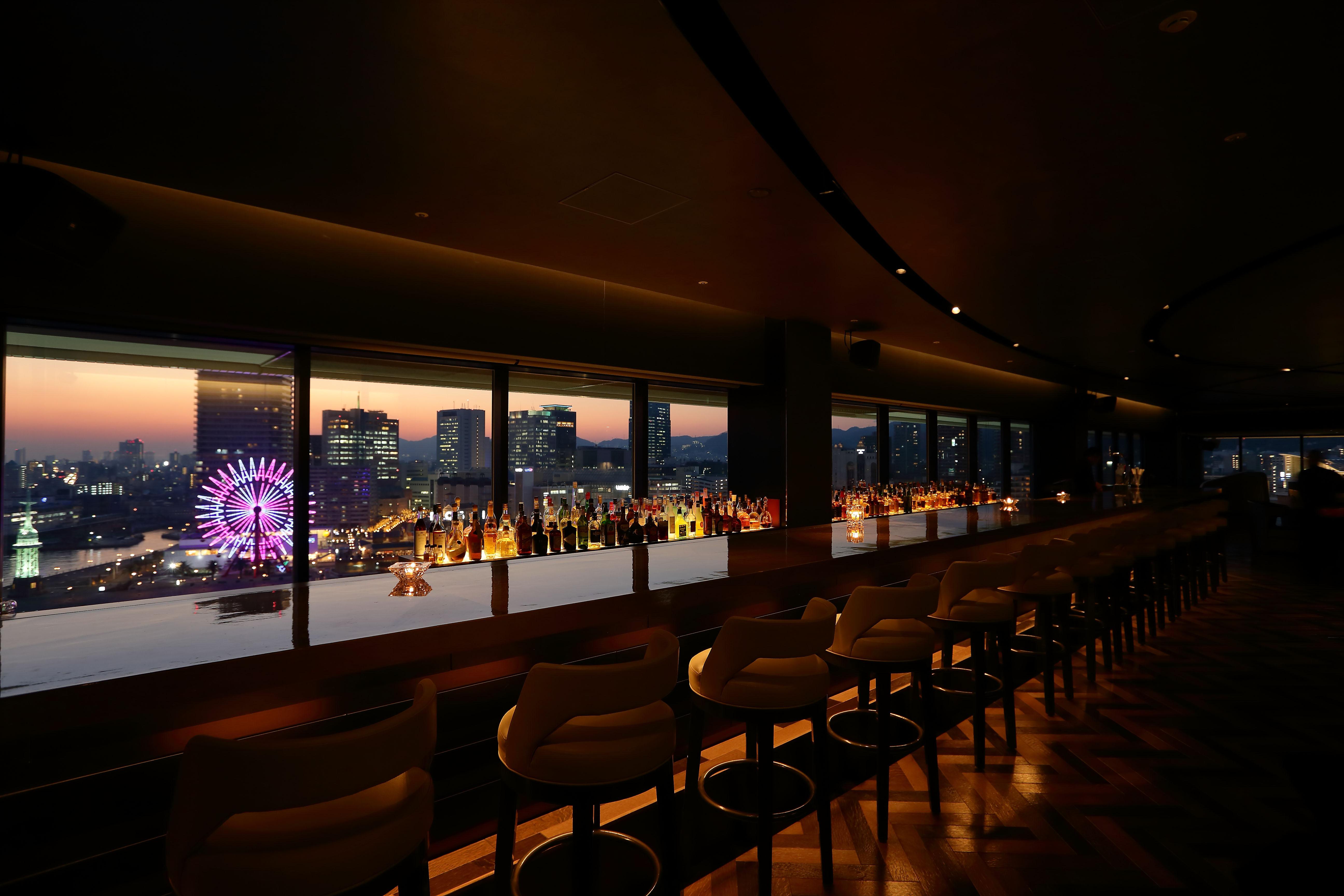 فندق كوبي ميريكين بارك أورينتال كوبه المظهر الخارجي الصورة The photo shows the interior of a stylish bar or lounge with a panoramic view of a city skyline at dusk. The lighting is dim, creating an intimate atmosphere. There are elegant bar stools lined up along a long counter, which features an array of illu
