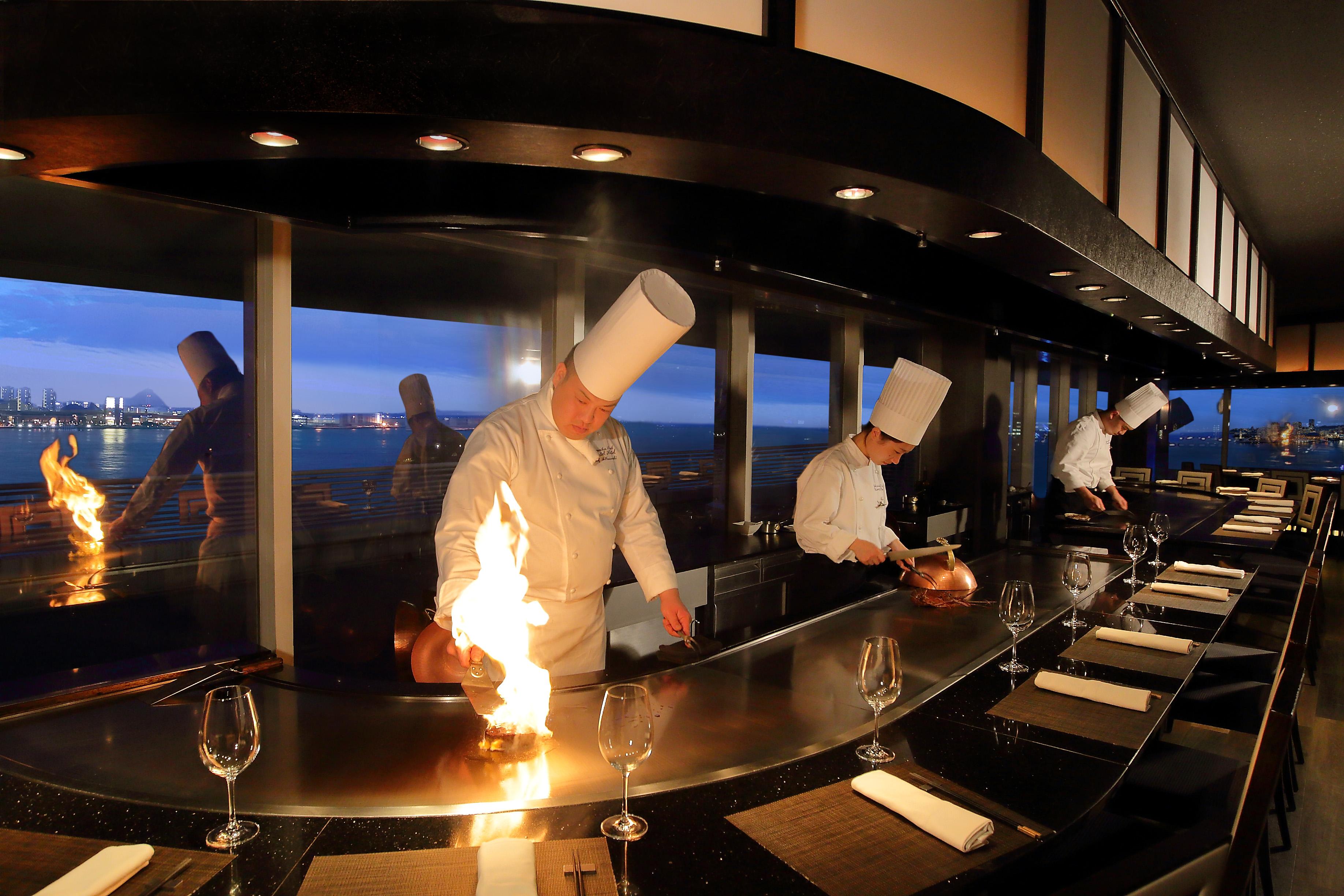 فندق كوبي ميريكين بارك أورينتال كوبه المظهر الخارجي الصورة The photo shows a restaurant setting with chefs cooking in an open kitchen, likely in a teppanyaki style. There are three chefs, all wearing white uniforms and tall chef hats, actively engaged in preparing food, with one chef creating a flame on the 