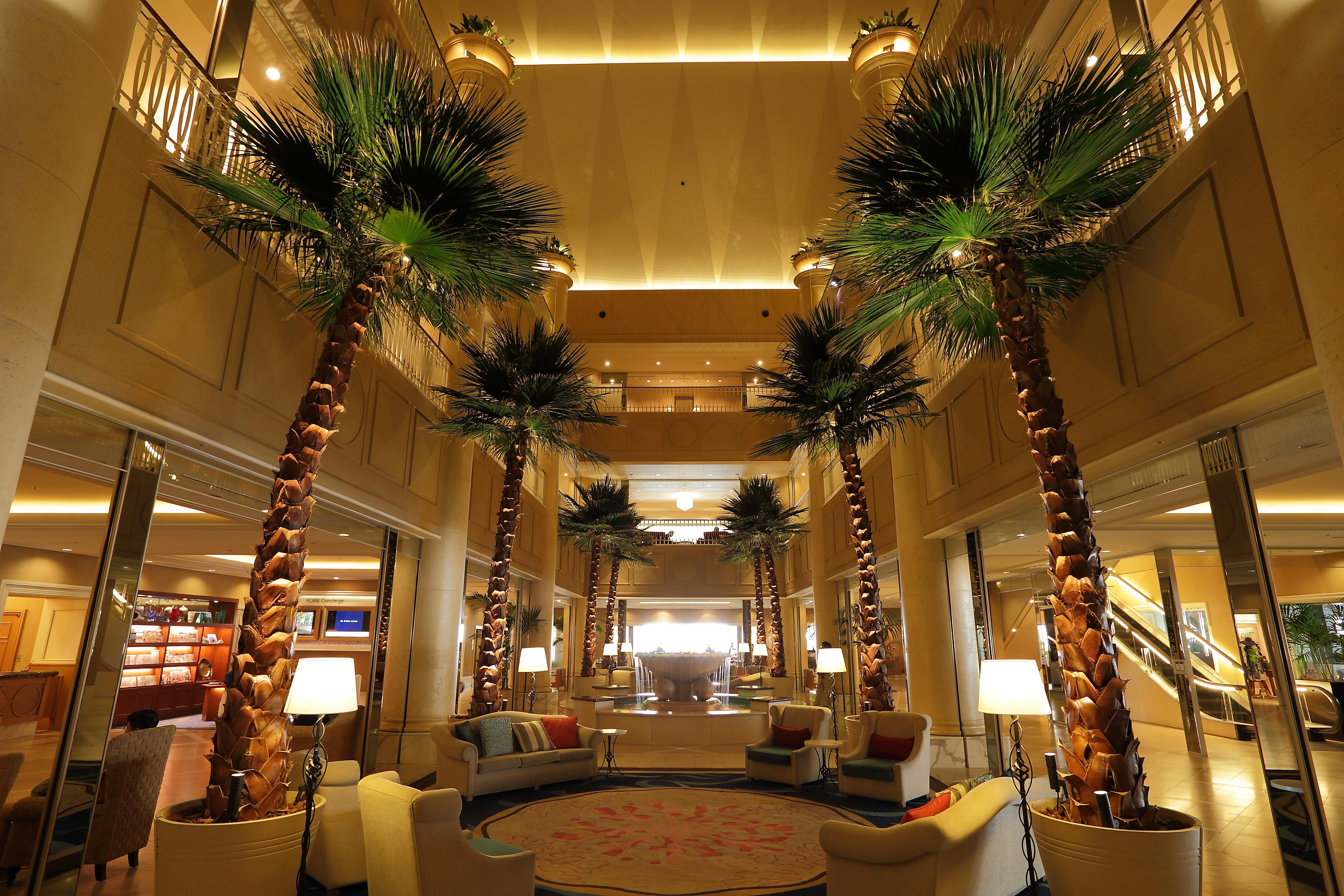 فندق كوبي ميريكين بارك أورينتال كوبه المظهر الخارجي الصورة The photo shows the interior of a luxurious hotel lobby. It features tall, elegant palm trees that create a tropical atmosphere. The space is well-lit, with a bright ceiling and warm lighting from decorative lamps. Comfortable seating arrangements, i