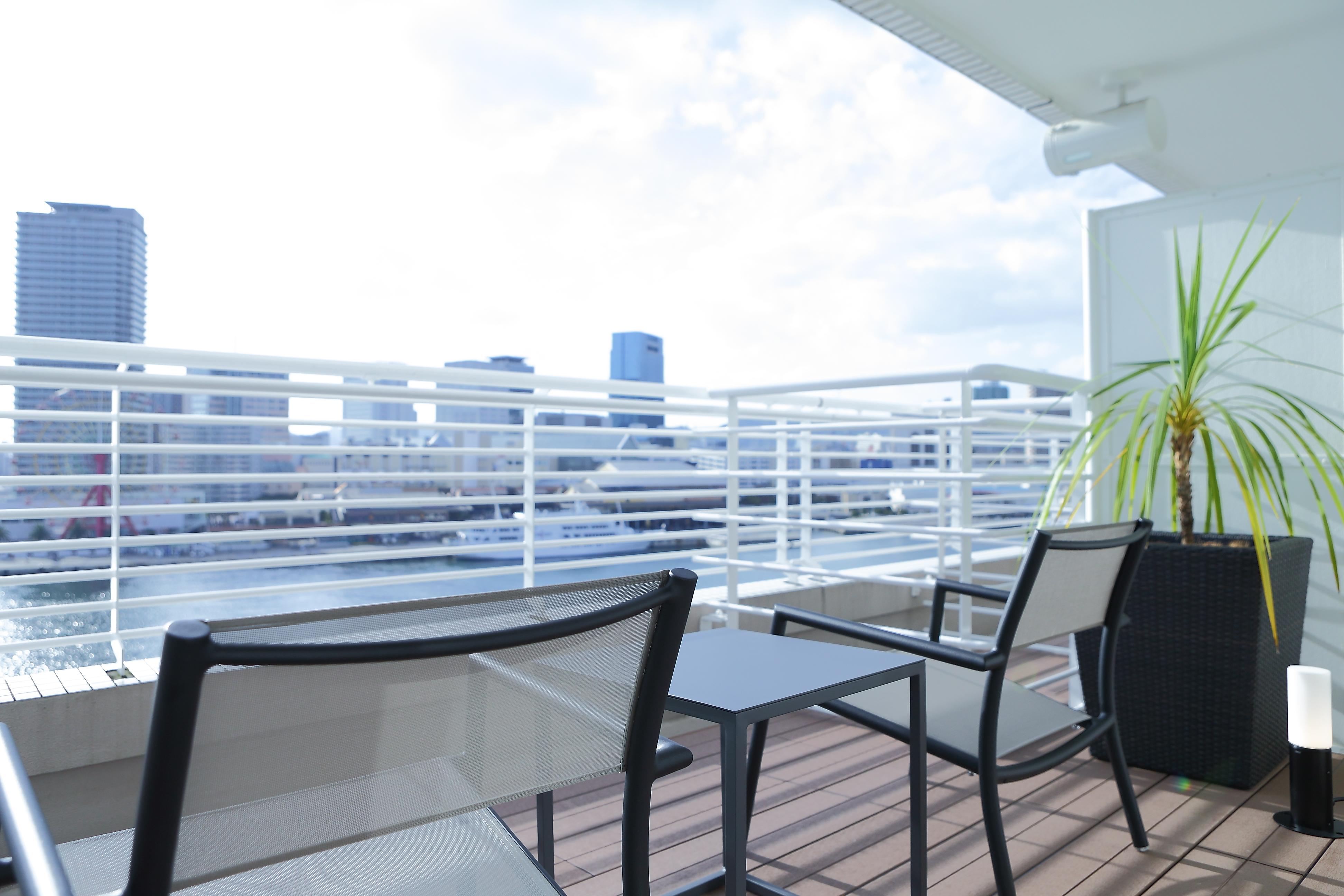 فندق كوبي ميريكين بارك أورينتال كوبه المظهر الخارجي الصورة The photo shows a modern balcony or terrace space with a view of a city skyline. There are two black chairs and a small table arranged on a wooden deck. In the background, there are several tall buildings visible, and the sky is partly cloudy. There 