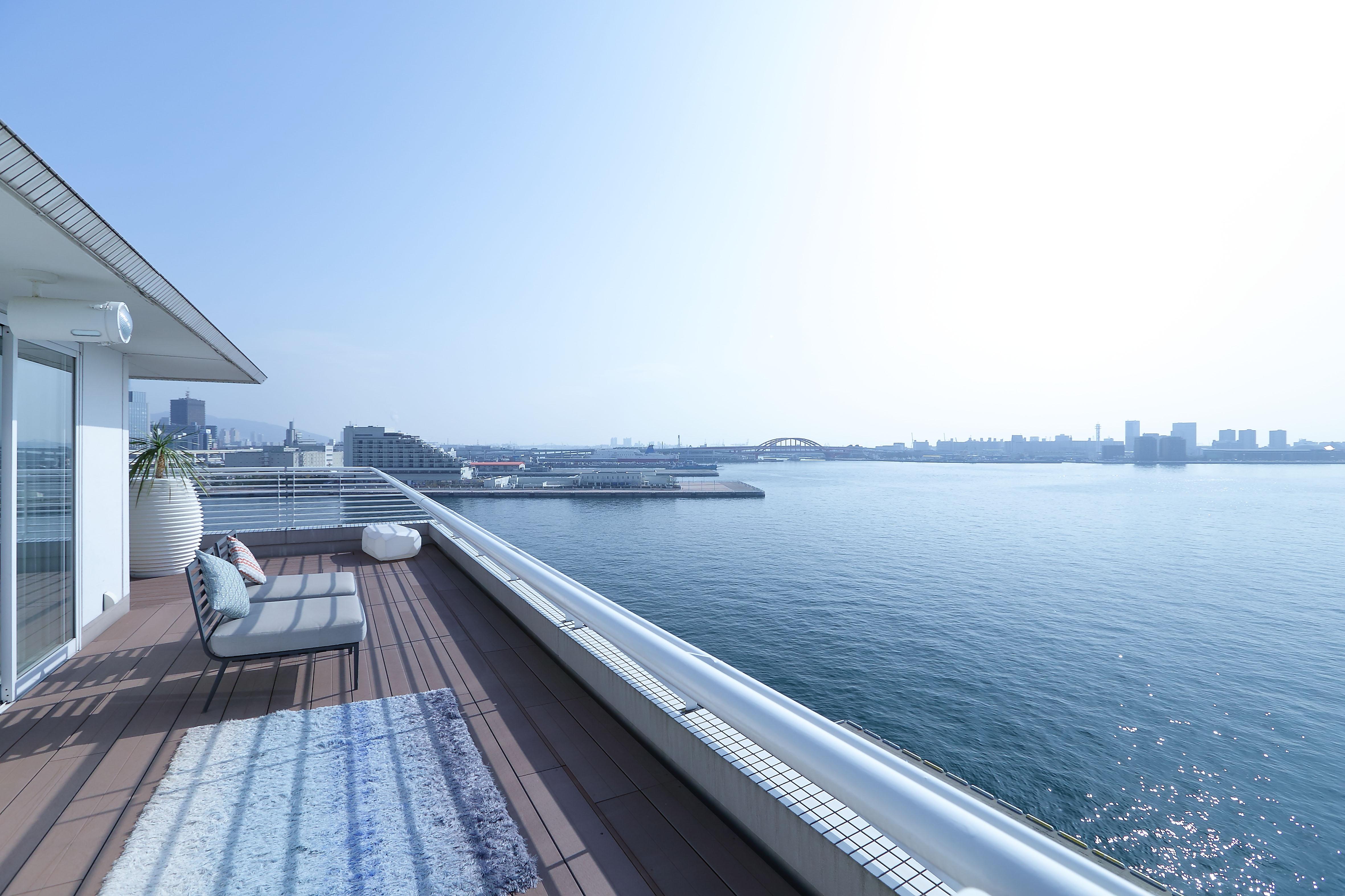 فندق كوبي ميريكين بارك أورينتال كوبه المظهر الخارجي الصورة The image shows a spacious balcony or terrace overlooking a body of water, likely a harbor or river, with city buildings in the background. The area is bright and well-lit, indicating a clear, sunny day. There is a modern lounge chair or sofa on the 