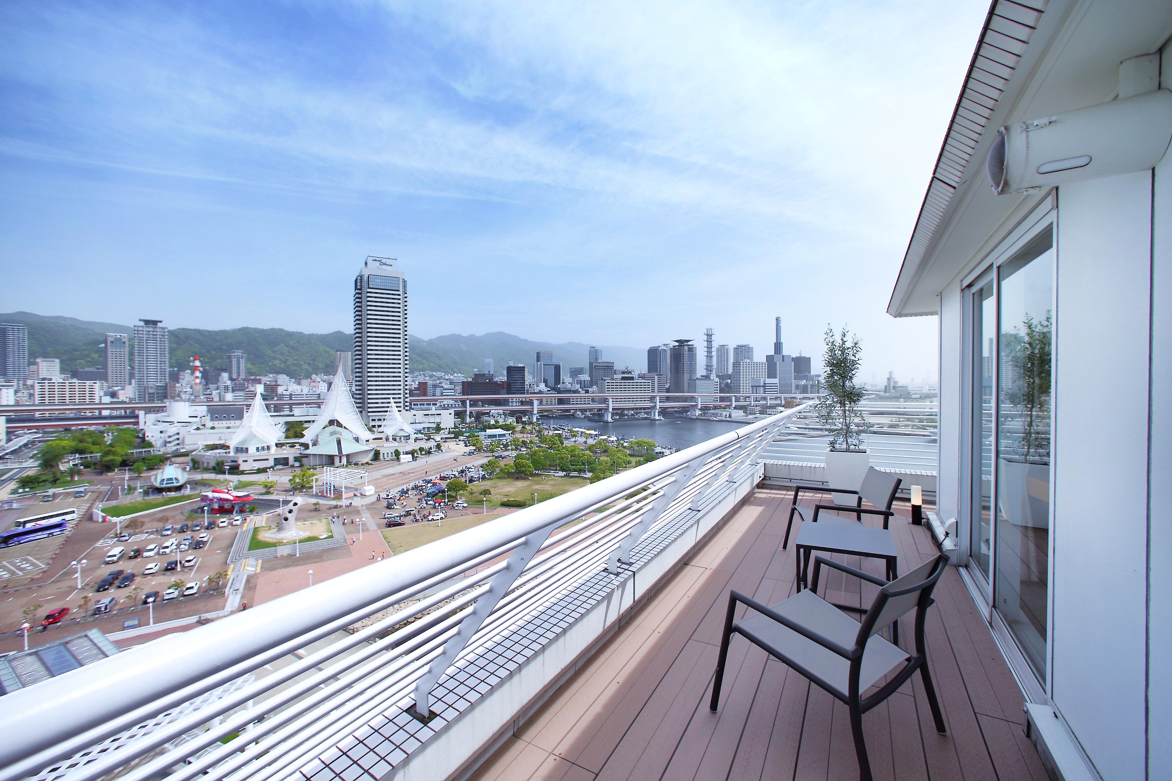 فندق كوبي ميريكين بارك أورينتال كوبه المظهر الخارجي الصورة The photo shows a balcony of a modern building with two chairs overlooking a cityscape. In the background, there are several high-rise buildings and a waterfront area. The scene is set against a clear sky with some clouds, and you can see greenery on