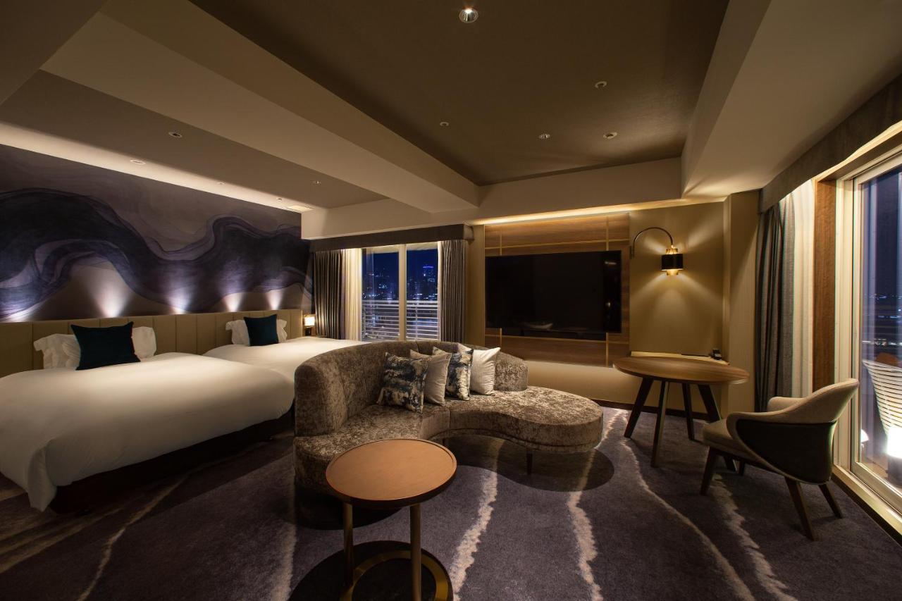 فندق كوبي ميريكين بارك أورينتال كوبه المظهر الخارجي الصورة The photo shows a modern hotel room with a stylish interior. There are two beds with white linens, and the decor features warm, ambient lighting. A plush, curved sofa sits in front of a round coffee table, and there’s a chair nearby. A large televisi