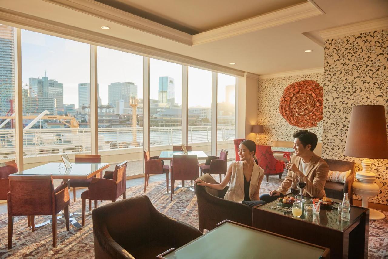 فندق كوبي ميريكين بارك أورينتال كوبه المظهر الخارجي الصورة The photo shows a modern, elegantly designed lounge area with large windows providing a view of a cityscape, including buildings by a river. Two people are seated in a cozy setting, enjoying each other's company. The interior features comfortable cha
