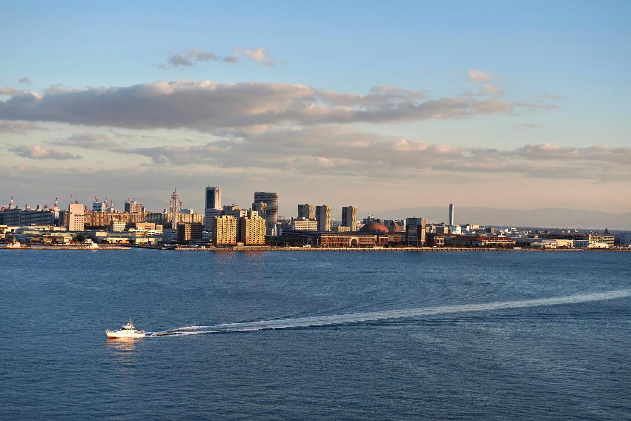 فندق كوبي ميريكين بارك أورينتال كوبه المظهر الخارجي الصورة The photo shows a calm waterfront scene with a city skyline in the background. The skyline features various tall buildings and structures, indicating an urban area. In the foreground, there is a boat on the water, leaving a wake as it moves. The sky 