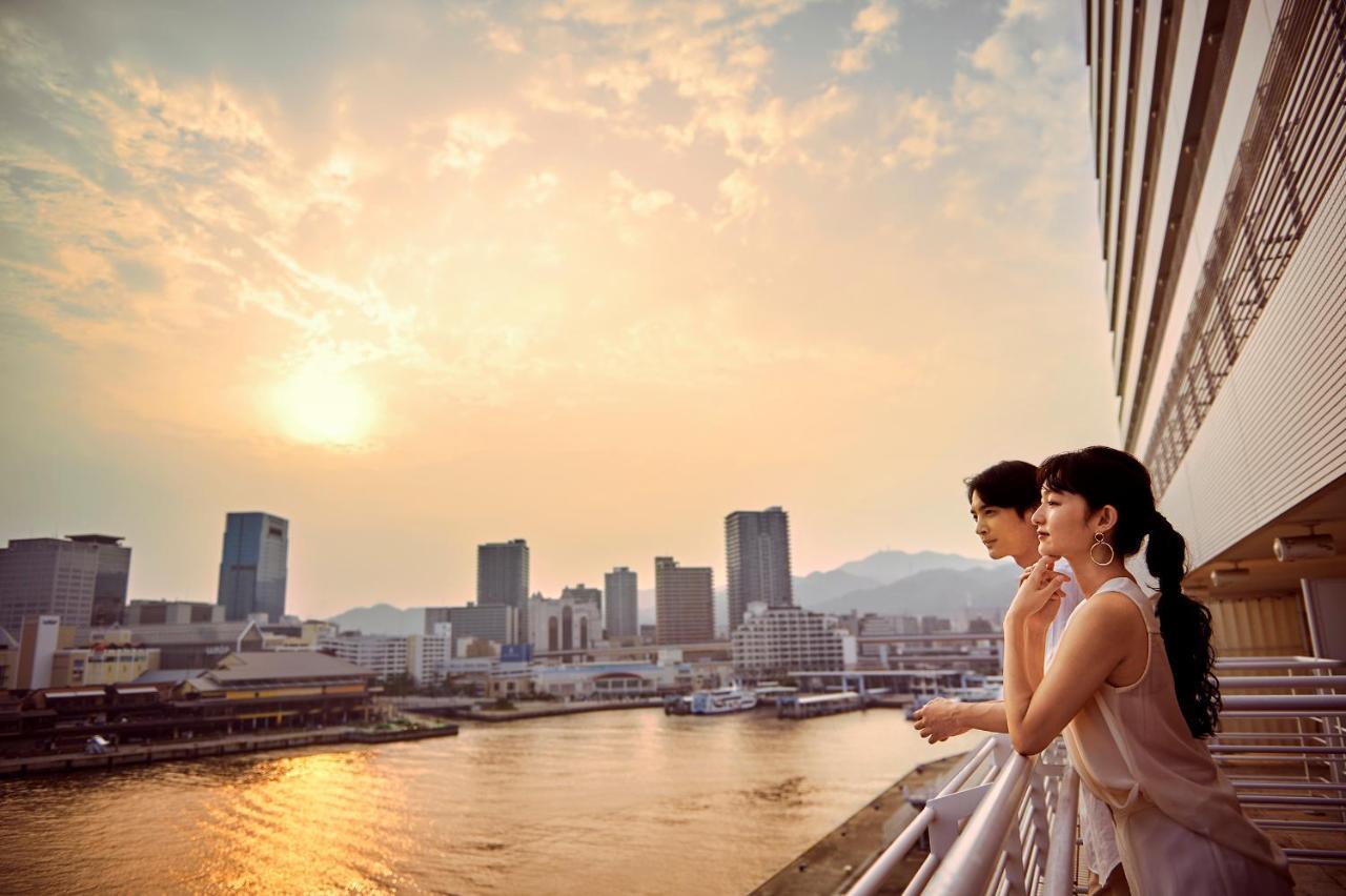فندق كوبي ميريكين بارك أورينتال كوبه المظهر الخارجي الصورة The photo depicts two people standing on a balcony, gazing out over a cityscape illuminated by a warm sunset. The scene captures a river reflecting the golden hues of the sky, with various buildings lining the waterfront. In the background, mountains