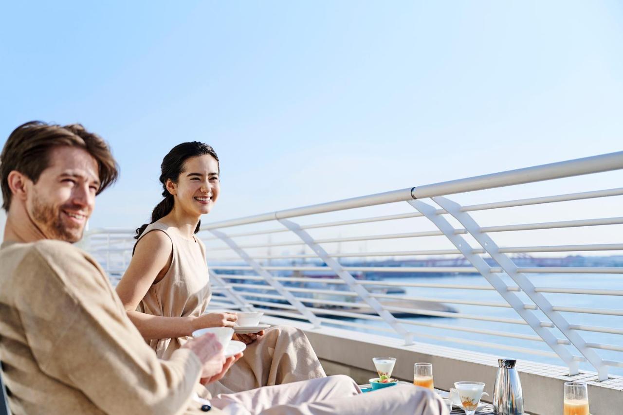 فندق كوبي ميريكين بارك أورينتال كوبه المظهر الخارجي الصورة The photo shows two people enjoying a pleasant moment outdoors, seated on a balcony or terrace. The scene has a bright, sunny atmosphere with blue skies and water in the background. The man has a casual look with a light-colored outfit, while the wom