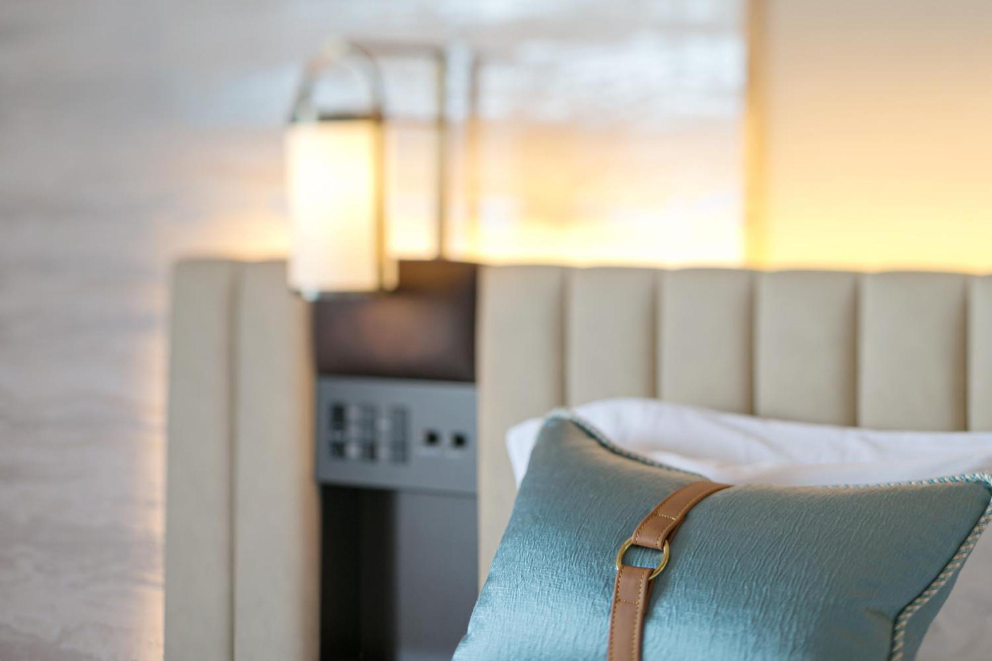فندق كوبي ميريكين بارك أورينتال كوبه المظهر الخارجي الصورة The photo shows a cozy and stylish bedroom setting. In the foreground, there is a decorative pillow with a blue fabric and a leather accent, resting on a beige upholstered bed. Behind the bed, there is a softly lit ambiance with a lamp or wall sconce