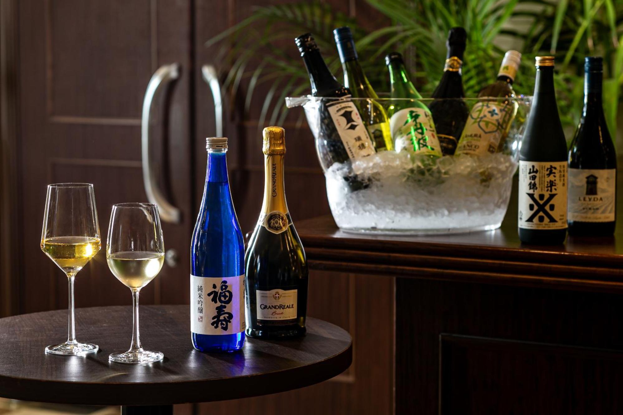 فندق كوبي ميريكين بارك أورينتال كوبه المظهر الخارجي الصورة The photo features a selection of alcoholic beverages displayed elegantly. On a dark wooden table, there are two wine glasses filled with a light-colored liquid, possibly white wine. Nearby, there is a blue bottle labeled with Japanese characters, al