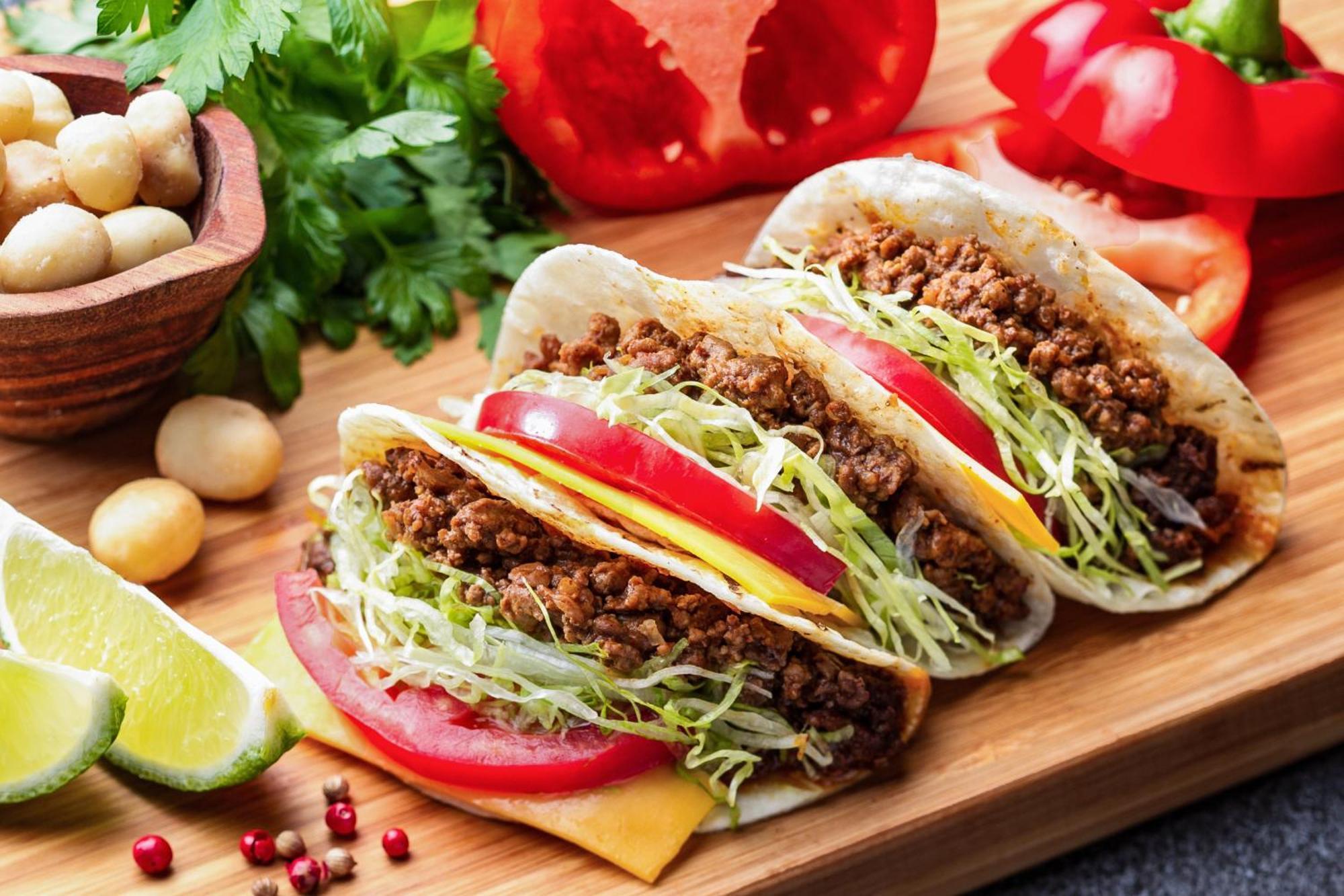 فندق كوبي ميريكين بارك أورينتال كوبه المظهر الخارجي الصورة The photo shows three tacos placed on a wooden serving board. Each taco is filled with seasoned ground meat, topped with shredded lettuce and slices of tomato. There are also slices of cheese visible inside the tacos. Surrounding the tacos are fresh 