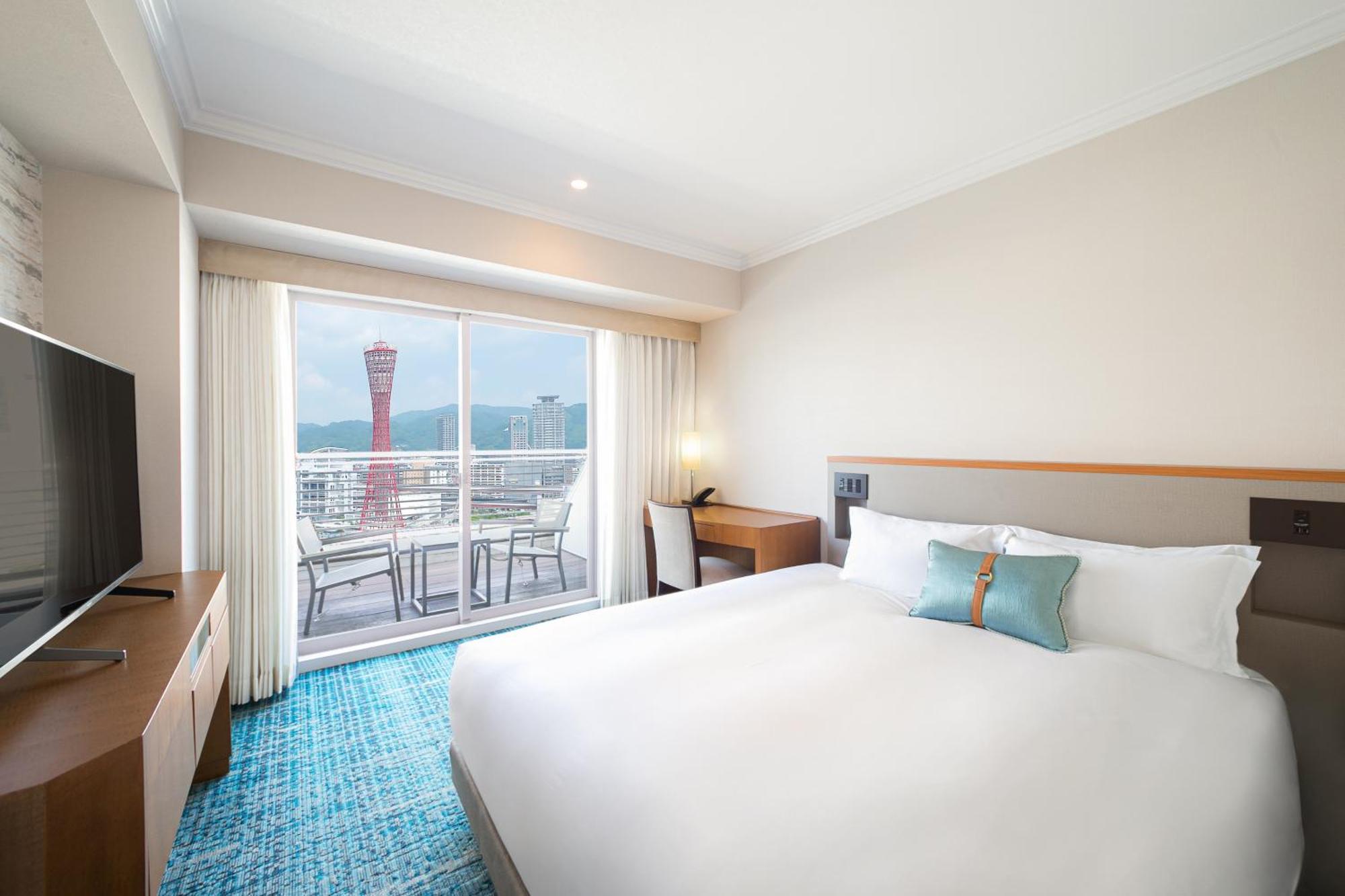 فندق كوبي ميريكين بارك أورينتال كوبه المظهر الخارجي الصورة The photo shows a modern hotel room with a large bed featuring white bedding and a teal accent pillow. There's a wooden desk and a flat-screen TV opposite the bed. Large windows provide a view of an outdoor area, which includes a balcony with furnitu