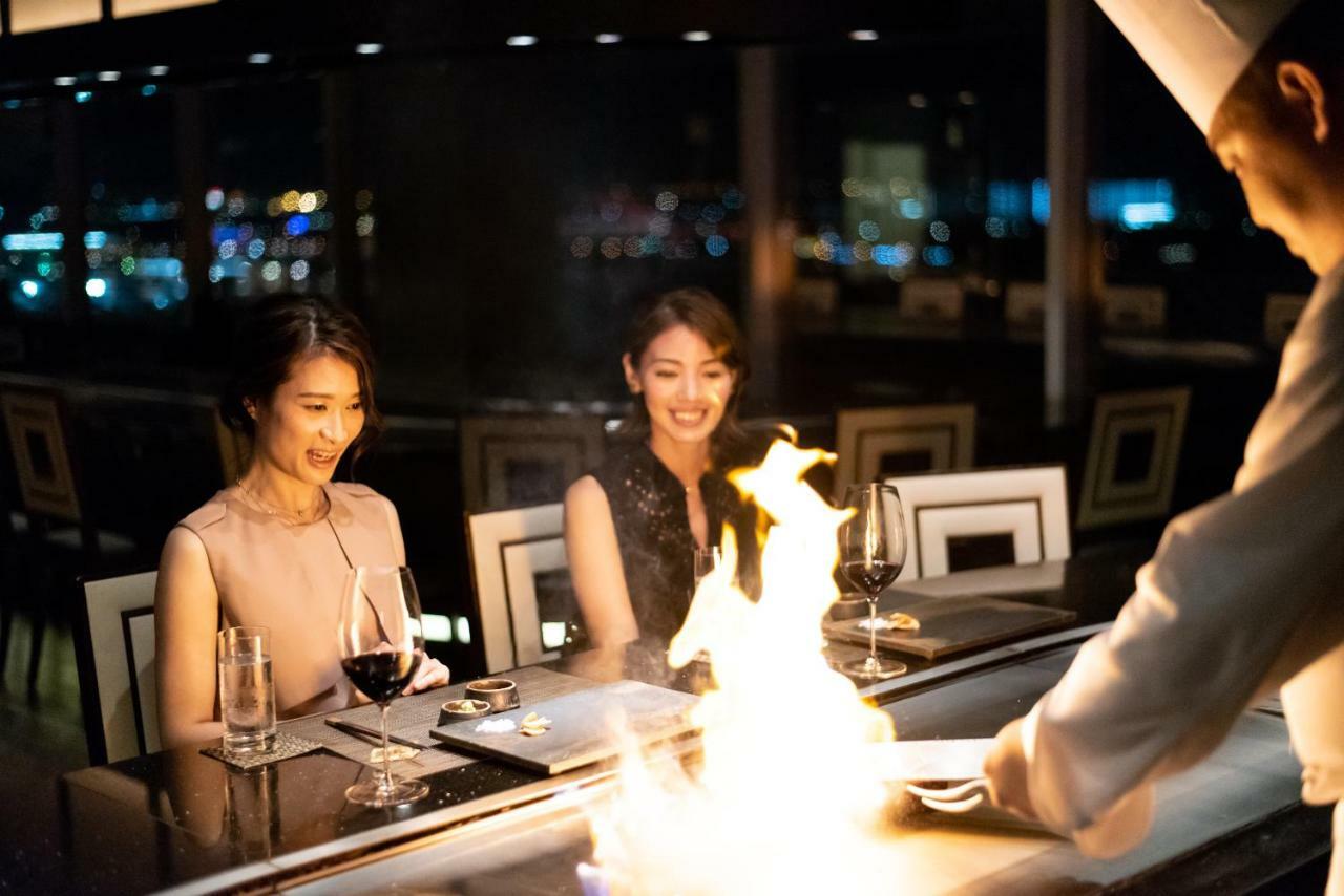 فندق كوبي ميريكين بارك أورينتال كوبه المظهر الخارجي الصورة The photo depicts a dining scene in a luxurious restaurant. Two women are seated at a table, enjoying the experience. They appear to be engaged and delighted by the culinary performance taking place in front of them. A chef is preparing food on a gri
