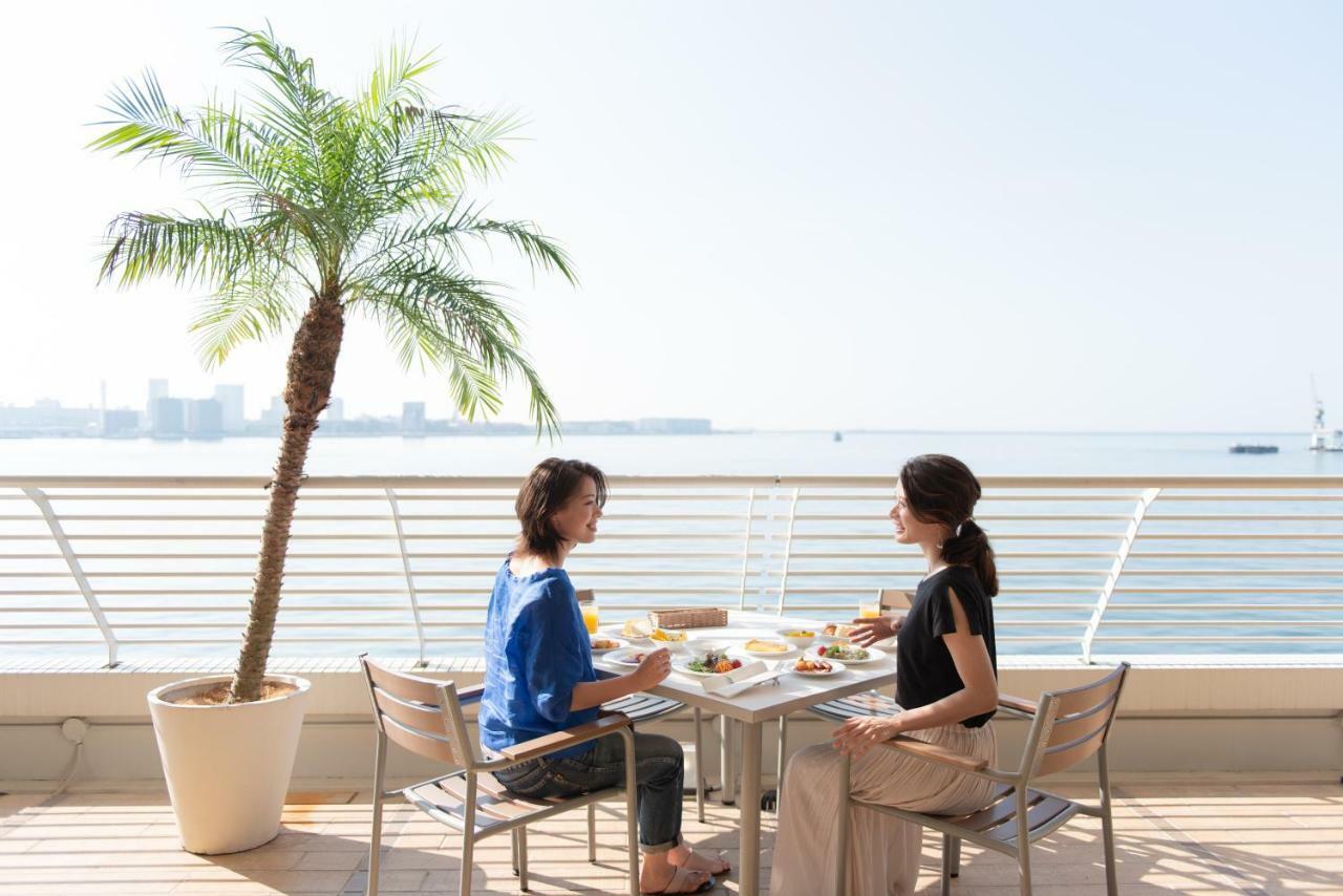 فندق كوبي ميريكين بارك أورينتال كوبه المظهر الخارجي الصورة The photo shows two women sitting at a table on a sunny terrace by the water. They are enjoying a meal together, with dishes laid out on the table in front of them. There is a tall palm tree in a planter nearby, and the view overlooks a body of water