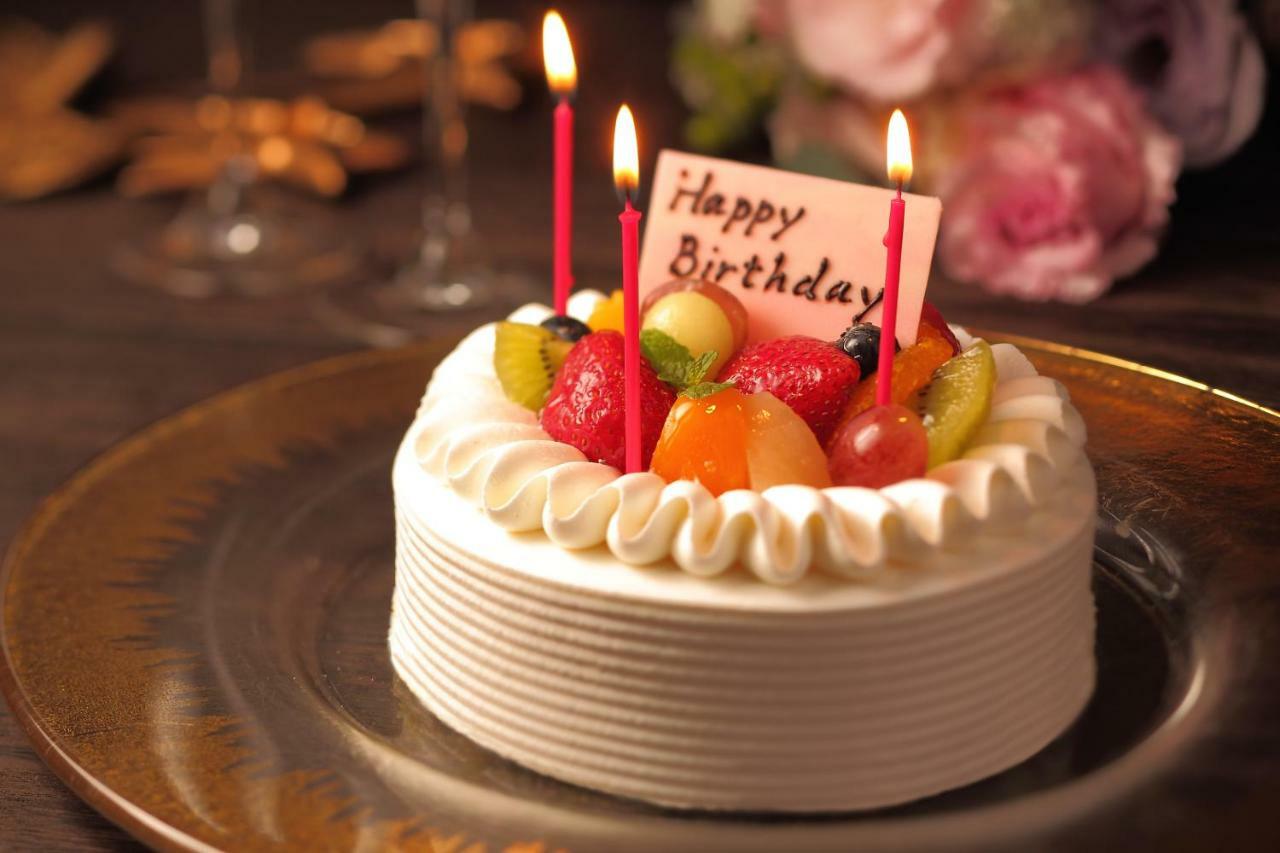 فندق كوبي ميريكين بارك أورينتال كوبه المظهر الخارجي الصورة The photo shows a beautifully decorated birthday cake placed on a plate. The cake is round with white frosting, and it features a variety of fresh fruits on top, including strawberries, grapes, and kiwis. There are four red candles lit on the cake, a